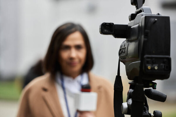 A multiracial female journalist wears a beige coat, confidently addressing the camera with a microphone in hand.