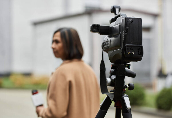 A multiracial female journalist in a beige coat prepares to deliver a news report in a bustling city atmosphere.