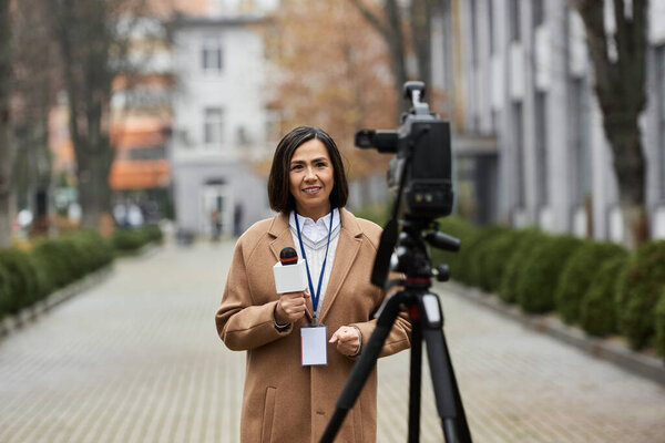 A confident journalist stands outdoors in a beige coat, holding a microphone and preparing to report news.