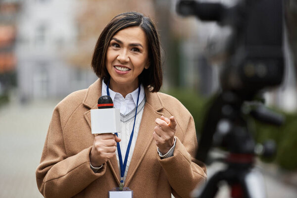A multiracial female journalist in a beige coat is actively engaging while holding a microphone outdoors.