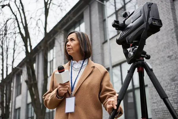 Bej ceketli çok ırklı bir kadın gazeteci bir mikrofon tutuyor ve açık hava haberlerine hazırlanıyor..