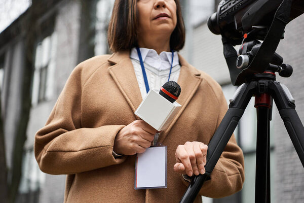 A multiracial female journalist in a beige coat stands ready with a microphone, preparing for an important report.