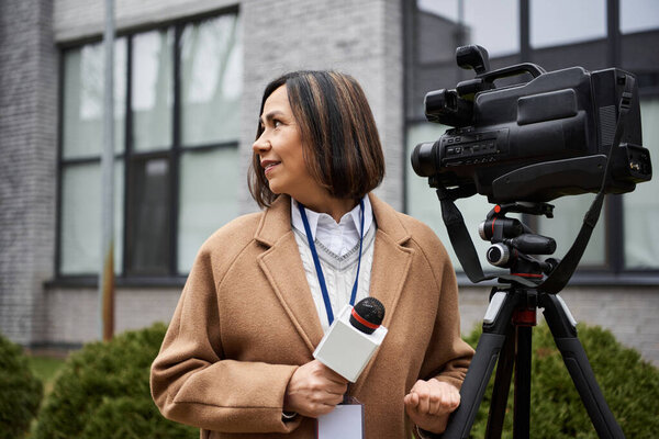 A journalist in a beige coat holds a microphone while preparing to report outside a modern building.