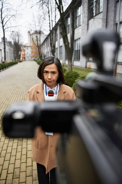 Многорасовая журналистка в бежевом пальто уверенно стоит, с микрофоном в руке, готовая поделиться новостями.