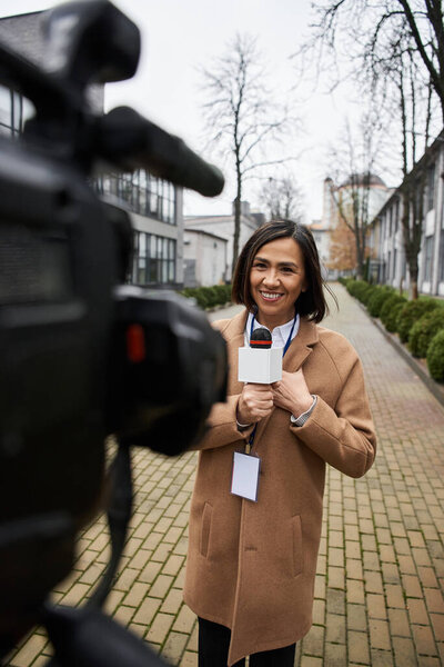 A multiracial female journalist stands confidently outdoors, holding a microphone, ready to report news.