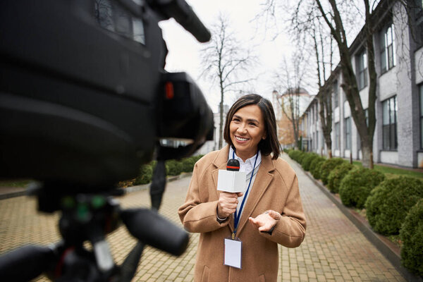 A multiracial female journalist holds a microphone, sharing lively news updates outdoors in an urban setting.
