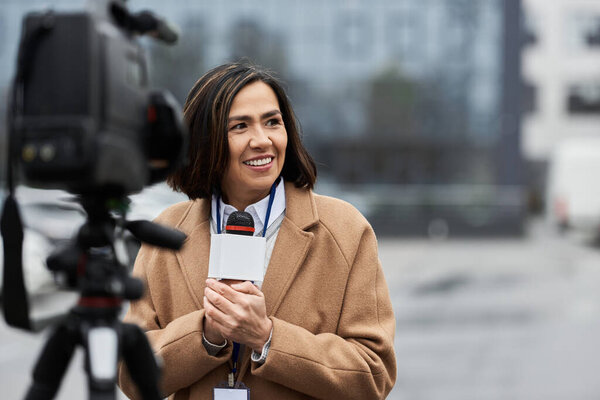 A smiling journalist holds her microphone, ready to deliver news updates outside in a vibrant city setting.