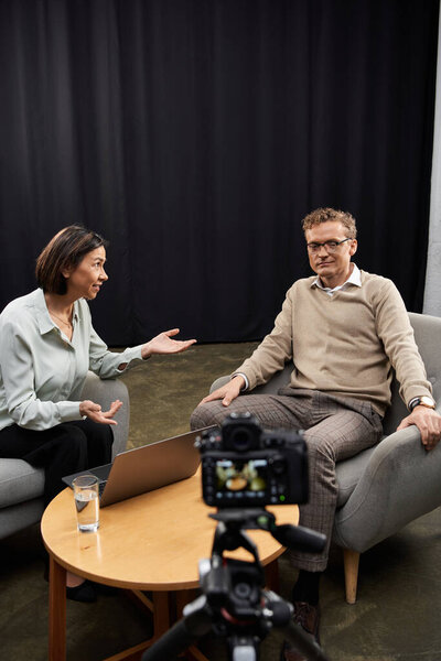 A middle-aged female journalist conducts a professional interview with a man seated on a stylish sofa.