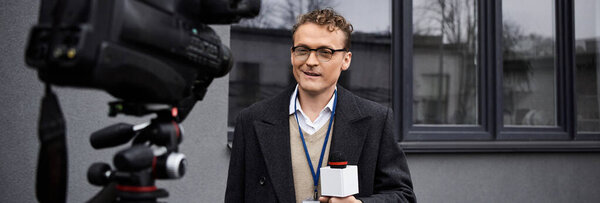 A journalist wearing glasses and a coat speaks into a microphone, sharing important news updates outside.