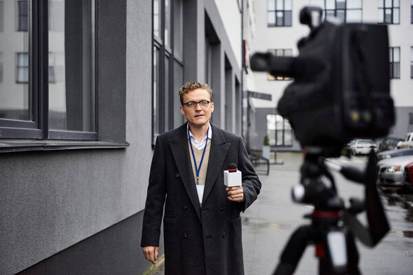A dedicated news anchor interviews outside, holding a microphone while wearing a coat and glasses.
