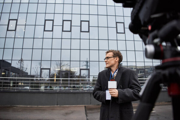 Ведущий новостей в очках с микрофоном в руке рядом с гладкой городской обстановкой.