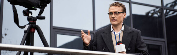 Сфокусированный журналист в пальто говорит в микрофон за пределами современной структуры в вечернее время.