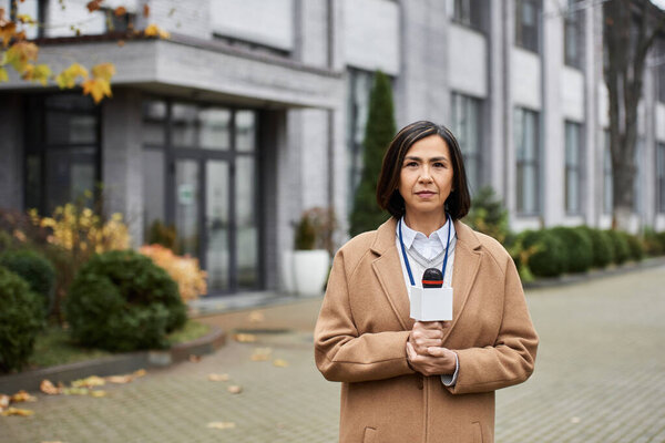 Многорасовая женщина в бежевом пальто уверенно стоит с микрофоном, сообщая на месте.