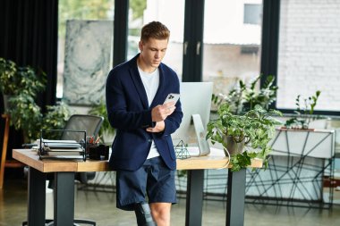 A confident young businessman with an artificial limb uses his smartphone while standing at his desk. clipart