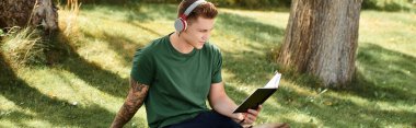Yakışıklı genç bir adam, kulaklık takıp, bir kitaba dalmış, dışarıda dinleniyor..
