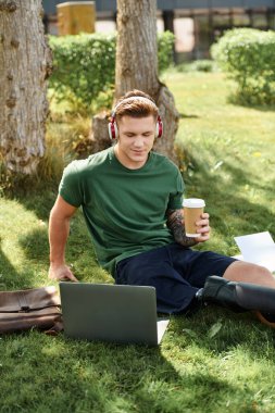 Protez bacaklı genç bir adam çimlerin üzerinde oturur ve laptopuyla çalışırken kahvesini yudumlar..