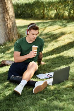 Yakışıklı genç bir adam çimlere oturur, dizüstü bilgisayarına odaklanır, kahvesini yudumlar, içeriği ve çeşitliliği kutlar..