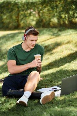 Protez bacaklı genç bir adam çimenlere oturur, içkisini yudumlar ve güneşli bir ortamda dizüstü bilgisayarına odaklanır..