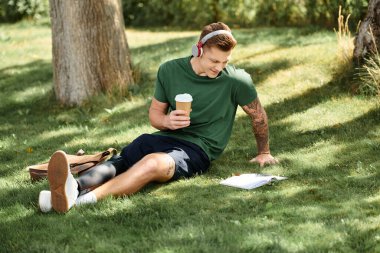 Yakışıklı genç bir adam çimenlerde rahatlar, kitap okurken kahvesini yudumlar, doğayı ve çeşitliliği kucaklar..
