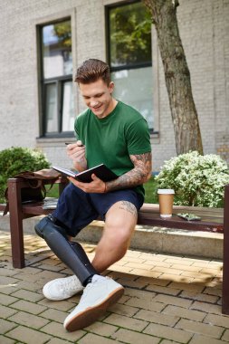 A young and handsome man with a prosthetic leg sits on a bench, smiling as he writes in a notebook. clipart