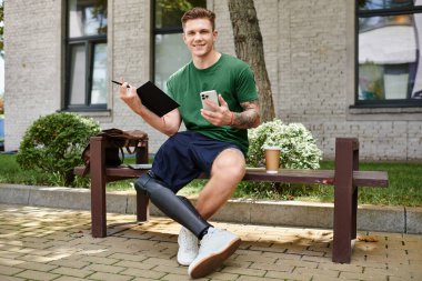 Dışarıda bir bankta oturan genç bir adam, bir defter ve telefonu dengede tutarken gülümsüyor, neşe ve yaratıcılığını ifade ediyor..