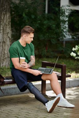 Protez bacağı olan genç bir adam bankta rahatlar, kahvesini yudumlar ve parktaki dizüstü bilgisayarına odaklanır..