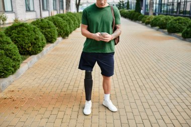 Protez bacaklı genç ve yakışıklı bir adam dışarıda duruyor, akıllı telefonunu kontrol ediyor ve atmosferin tadını çıkarıyor..