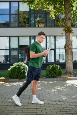 Protez bacaklı, genç, yakışıklı bir adam güvenli bir şekilde dışarıda yürüyor, akıllı telefonuna odaklanıyor..