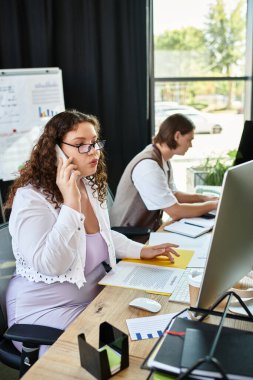 A young plus size woman speaks on the phone while her male friend works diligently at a desk. clipart