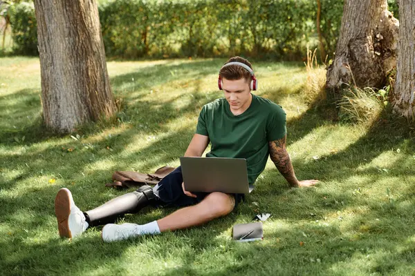 Genç bir adam çimlerin üzerinde oturur, ağaçlarla çevrili bir dizüstü bilgisayar kullanır, çeşitliliği kararlılıkla ve dikkatle kutlar..