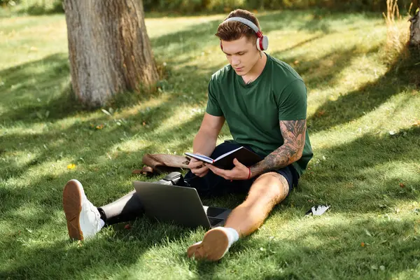 Protez bacaklı genç bir adam çimlerin üzerinde oturur, açık hava güneşinin tadını çıkarırken defterine odaklanır..