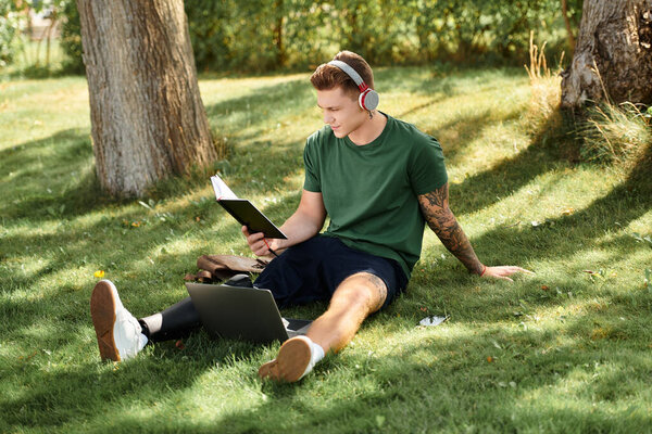 A young man relaxes on the grass, reading intently while wearing headphones and enjoying nature beauty.