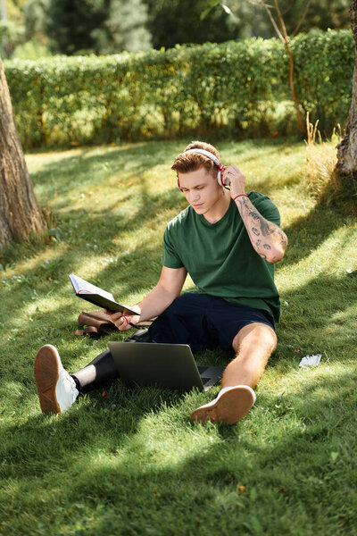 Красивый молодой человек с протезной ногой сидит на траве и учится, слушая музыку в парке.