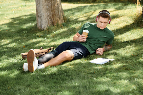 A young man relaxes on the grass with headphones, sipping coffee and reading his notes in a sunny park.