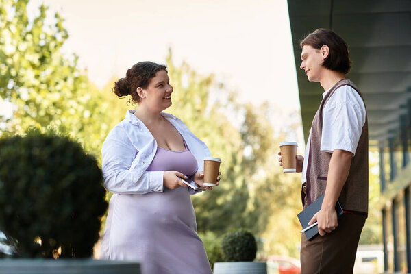 Молодая женщина плюс размер смеется со своим другом-мужчиной, держа кофейные чашки снаружи.