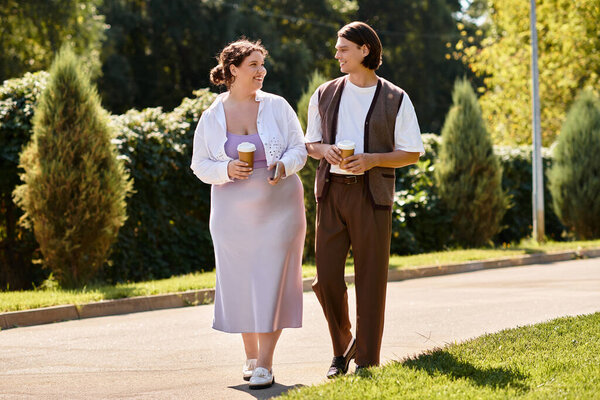 A young woman and her male friend stroll through a vibrant park, sipping coffee together.