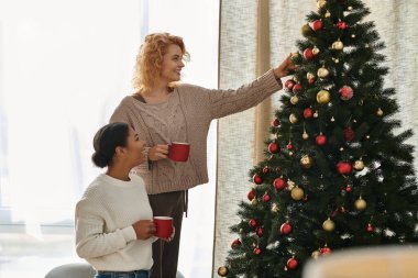 Two women share laughter and love while decorating a cheerful Christmas tree in their cozy home. clipart