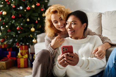 A couple enjoys cuddling together while looking at their phone, celebrating the holidays at home. clipart