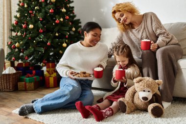 A lesbian couple shares festive treats and warm drinks with their daughter by the tree. clipart