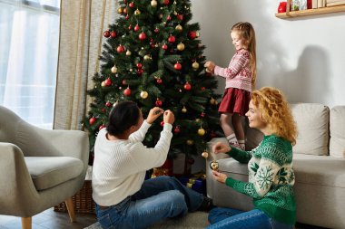 A loving lesbian couple and their daughter joyfully decorate a Christmas tree together. clipart