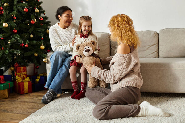 В уютной гостиной пара делится смехом со своей дочерью, пока она держит плюшевого мишку..