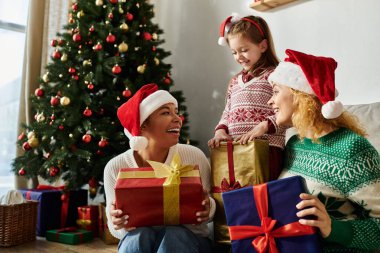 A happy lesbian couple and their daughter enjoy opening gifts together by the Christmas tree clipart