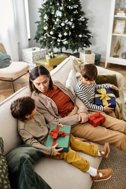 Bir anne, kışın parıldayan Noel ağacının hediyelerini açarken iki mutlu çocuğuyla oturur..