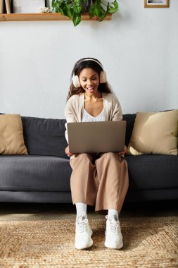 A young woman happily engages with her laptop while relaxing on a comfortable couch. clipart