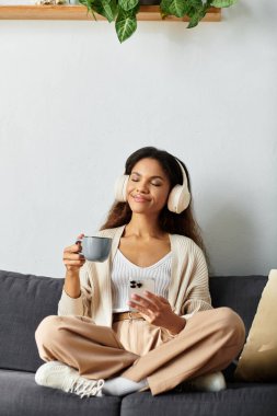 Neşeli bir kadın kanepede rahatça oturur, müzik dinlerken kahvenin tadını çıkarır..