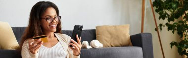 Neşeli bir kadın internetten alışveriş yapıyor, elinde bir kredi kartı ve akıllı telefon tutuyor, neşe saçıyor..