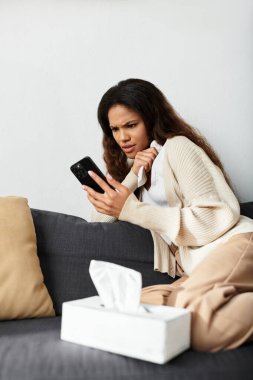 Bir kadın, akıllı telefonuna bakarken endişeli bir şekilde koltuğa rahatça oturur..