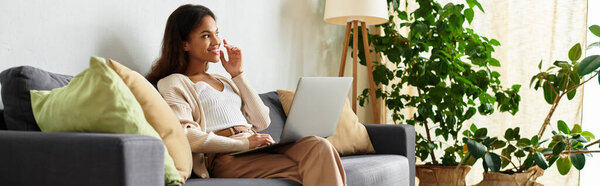 Женщина отдыхает на удобном диване, фокусируясь на ноутбуке посреди пышного внутреннего сада.