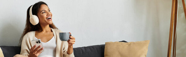 A cheerful young woman sips from a cup, immersed in music with headphones on, surrounded by comfort.