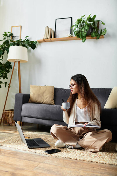 Sipping coffee, a woman sits cross legged on the floor, typing on her laptop in a serene space.